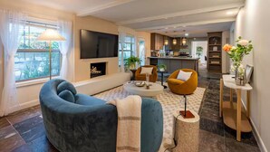 A modern living room with a large TV, comfortable seating, a fireplace, and a kitchen in the background. - Peacock Paradise - Pool / Game (Houston)