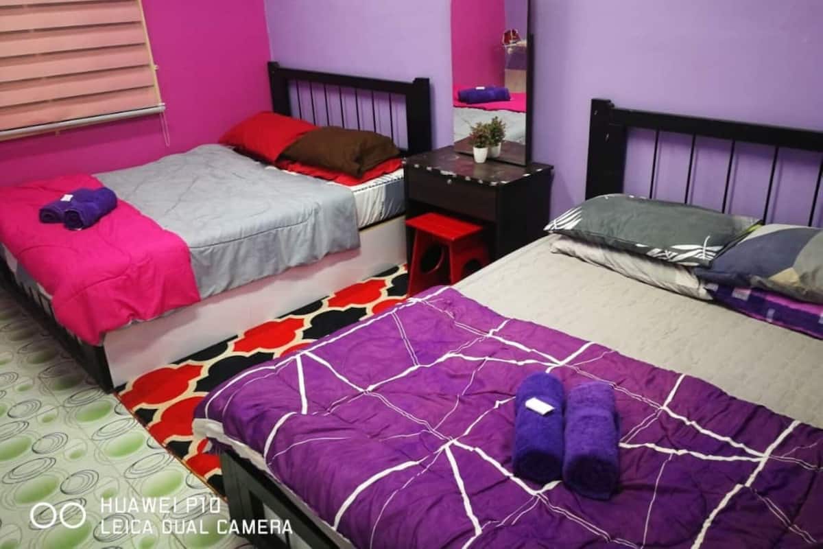 A room with two beds, one with a purple and white patterned bedspread and the other with a grey and red bedspread. There is a small desk with a mirror and a plant on it.