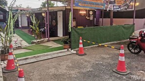 Entrada de hotel con una zona de espera techada, césped verde y conos de tráfico que señalan un área restringida.
