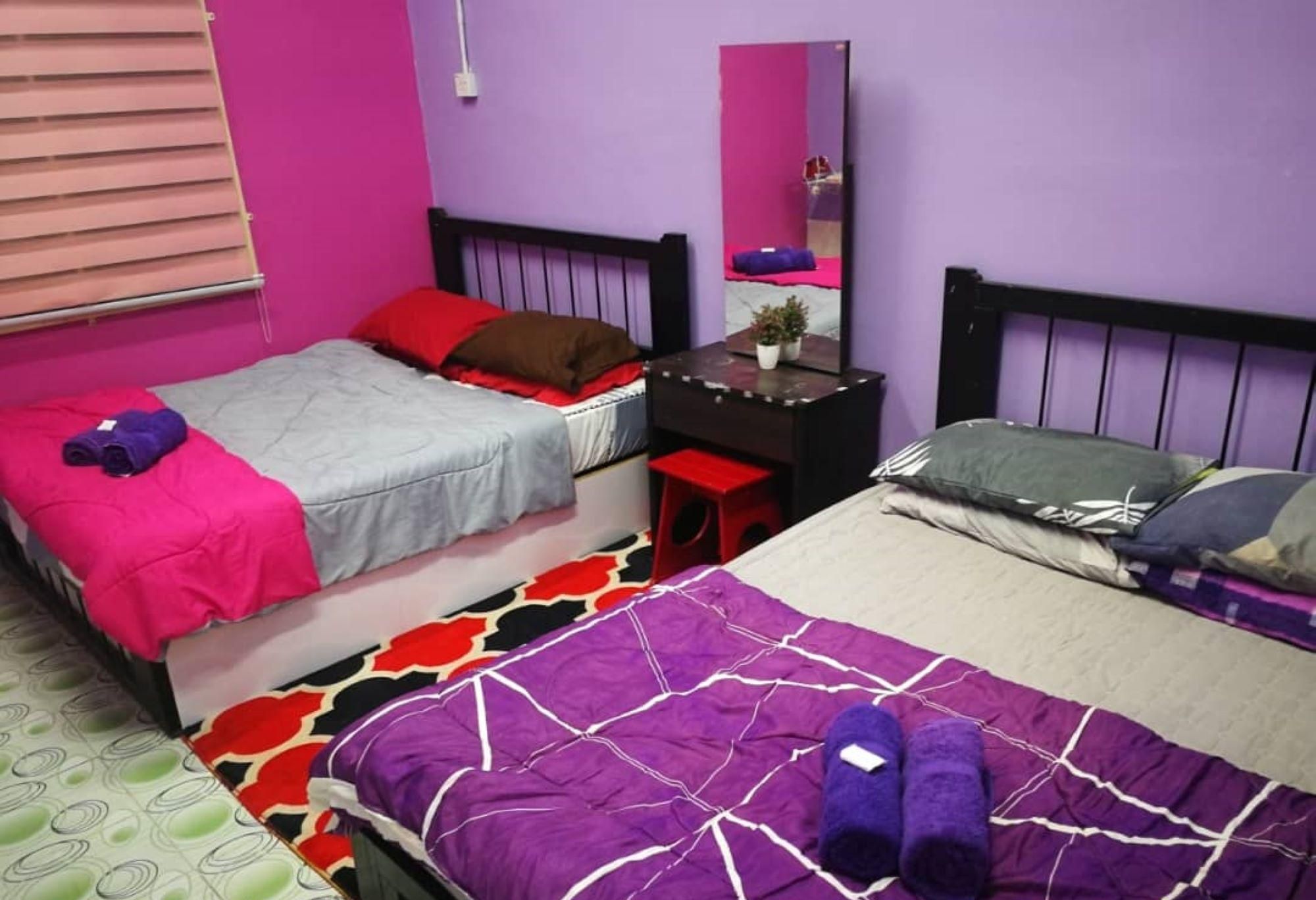 Una habitación con dos camas, una con colcha morada y blanca y la otra con cubrecama gris y rojo. Hay un escritorio pequeño con un espejo y una planta encima.
