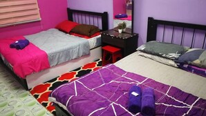 Une chambre avec deux lits : l’un est recouvert d’une couvre-lit à motifs violets et blancs, et l’autre d’une couvre-lit grise et rouge. On y trouve un petit bureau avec un miroir et une plante.