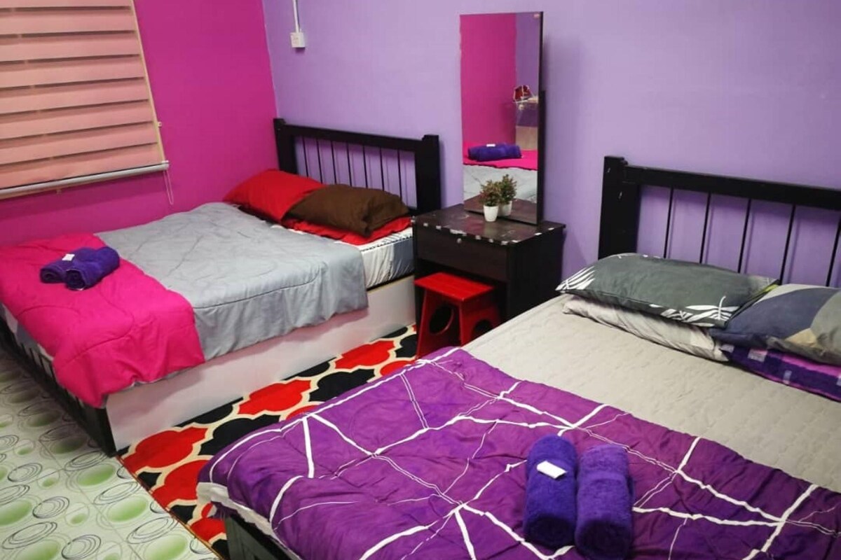 Een kamer met twee bedden, waarvan er één een paars-wit beddegoed heeft en de andere een grijs-rode spreiding. Er staat een klein bureau met een spiegel en een plant erop.