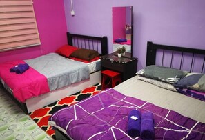 Een kamer met twee bedden, waarvan er één een paars-wit beddegoed heeft en de andere een grijs-rode spreiding. Er staat een klein bureau met een spiegel en een plant erop.