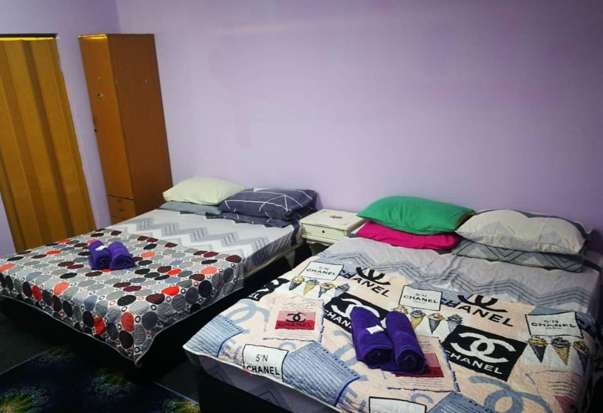 Habitación con dos camas, cada una con una colcha y cojines estampados. Se observa un armario de madera y una mesita de noche con una lámpara.