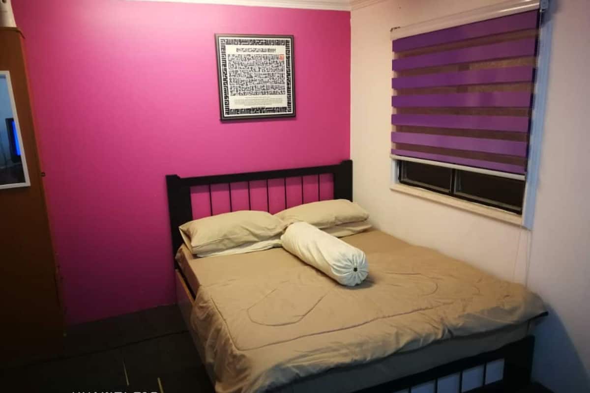A small bedroom with a pink wall, a bed with beige bedding, and a window with purple blinds.