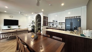 Una cocina moderna con armarios blancos, horno integrado y refrigerador. Hay un comedor con mesa y sillas de madera, y un televisor montado en la pared.