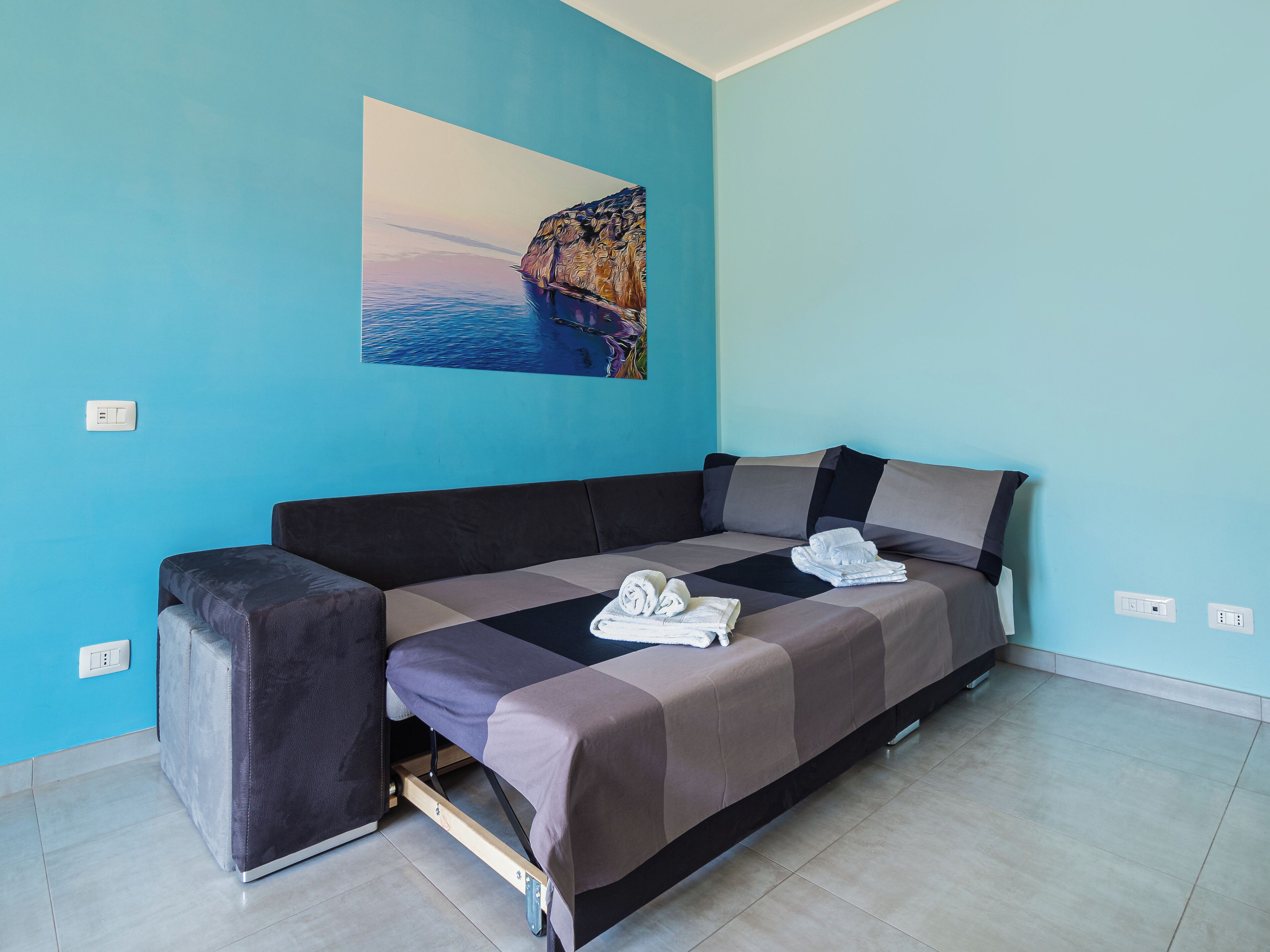 A room with a bed, a sofa, a painting, and a tiled floor.