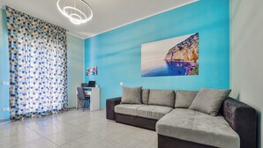 Ein modernes Wohnzimmer mit blauer Akzentwand, einem grauen Ecksofa, einem Schreibtisch mit Laptop und einem großen Gemälde.