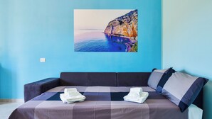 Ein modernes Zimmer mit blauer Wand, einem großen Gemälde einer Küstenlandschaft, einem dunklen Sofa mit kariertem Überwurf und ordentlich gefalteten Handtüchern auf der Sitzfläche.