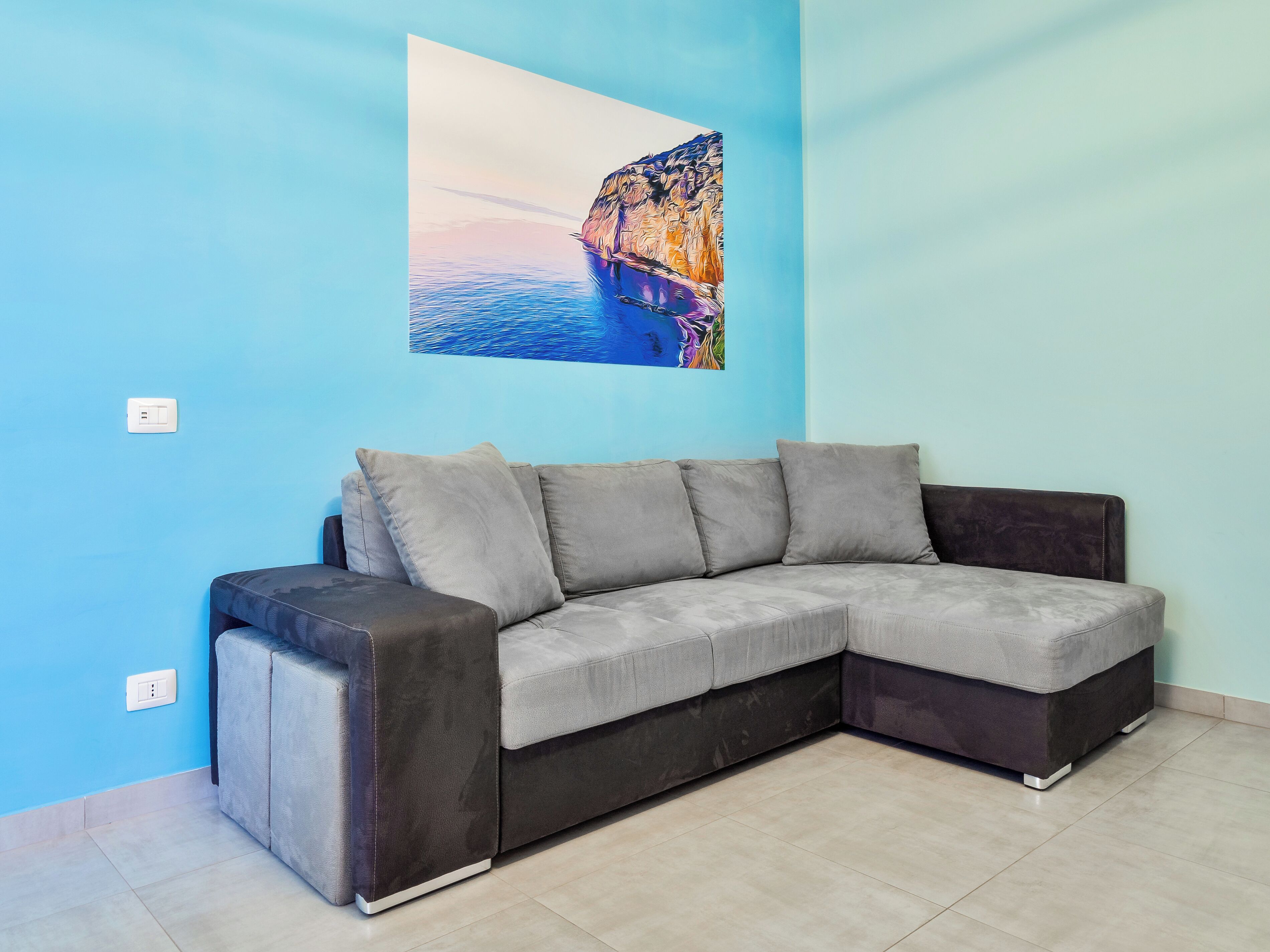 A corner living room with a grey sectional sofa, a blue wall, and a painting of a coastal scene.