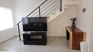 Uma sala de estar com uma TV de tela plana em um suporte preto, uma mesa consola de madeira, uma lixeira branca e uma escada.