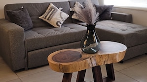 Uma sala de estar com um sofá cinza, uma mesa de centro redonda de madeira com um vaso, e uma janela com vista para árvores.