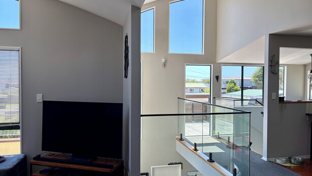 A modern living room with a flat-screen TV, wooden entertainment unit, and a staircase with glass railings leading to a balcony. - Beachlands Getaway - Room for everyone with a pool and views (Beachlands)