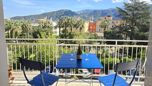 Un balcon avec une table et des chaises, donnant sur un paysage montagneux parsemé de bâtiments et de palmiers.