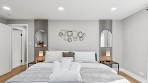 A modern hotel room with a large bed, two bedside tables with lamps, a wall-mounted mirror, and a decorative wall piece. - Comfortable Luxurious 4BR Home w/ Game Room + Master Suite (Mount Prospect)