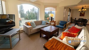 A spacious living room with a large window, a television, a sofa, a coffee table, and a dining area. - Slopeside Penthouse Apartment with Pool, Hot Tub and Sauna (Whistler)
