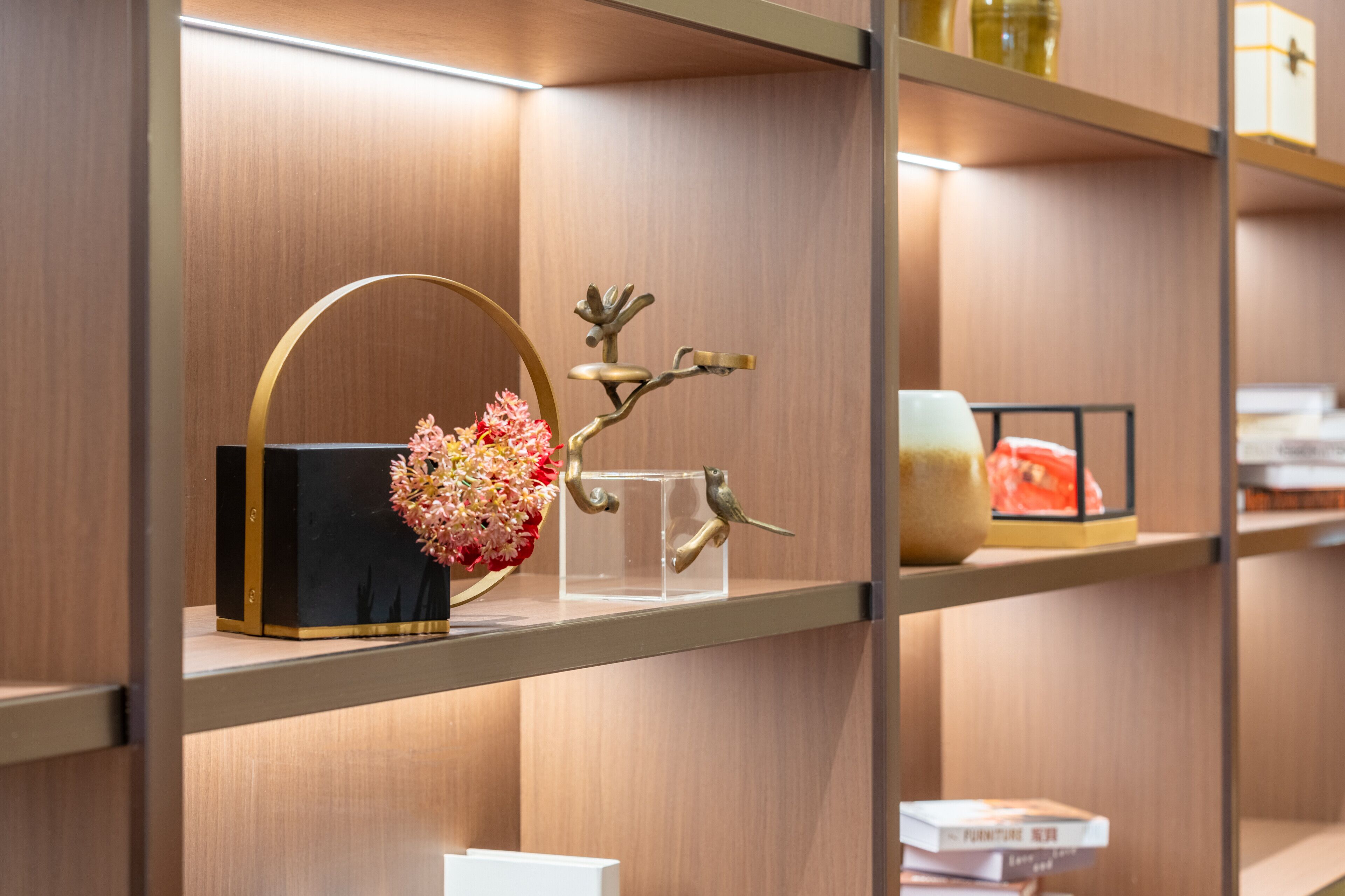 A shelf with a black vase with pink flowers, a clear glass with a gold bird, and a small white box.