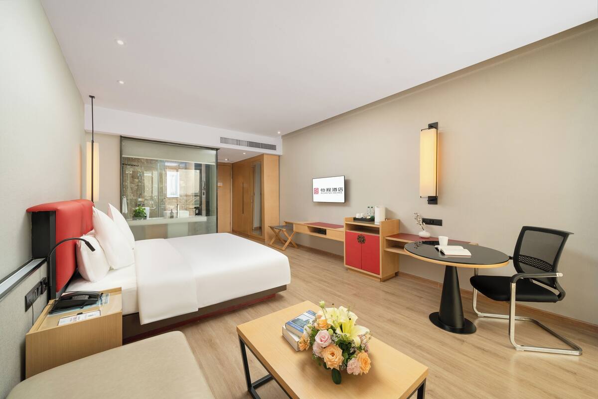 A modern hotel room with a bed, desk, chair, and a small table with a vase of flowers.