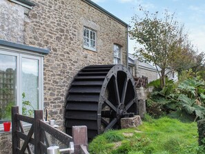 A stone building with a large water wheel, a wooden fence, and a tree. - The Cottage - UK50652 (Falmouth)