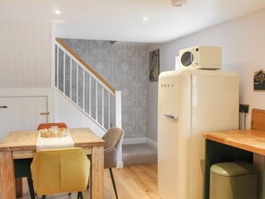A compact kitchen with a wooden table, a yellow chair, a white refrigerator, and a staircase leading to another room. - The Cottage - UK50652 (Falmouth)