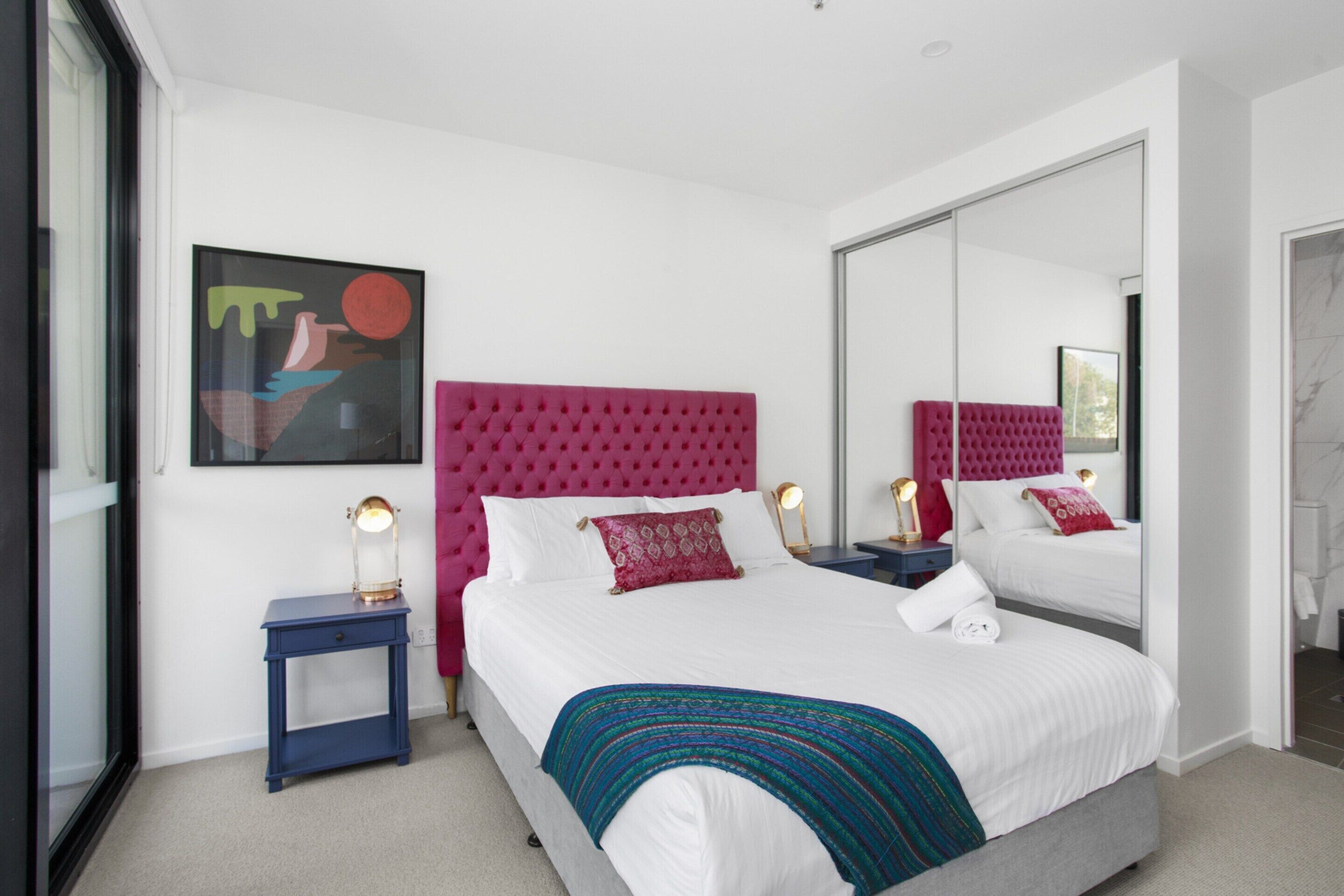 Une chambre moderne avec un grand lit, une tête de lit rose, une table de chevet avec une lampe et un grand miroir.