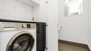 Ett tvättrum med en tvättmaskin och en torktumlare från Fisher & Paykel, en vit kakelvägg och en spegel som reflekterar rummet.