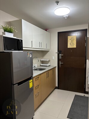 Eine kleine Küche mit weißen Schränken, einem Edelstahlkühlschrank, einer Mikrowelle und einem Spülbecken. Eine Holztür trägt ein Schild, und auf der Mikrowelle steht ein kleiner Topf mit Pflanze.