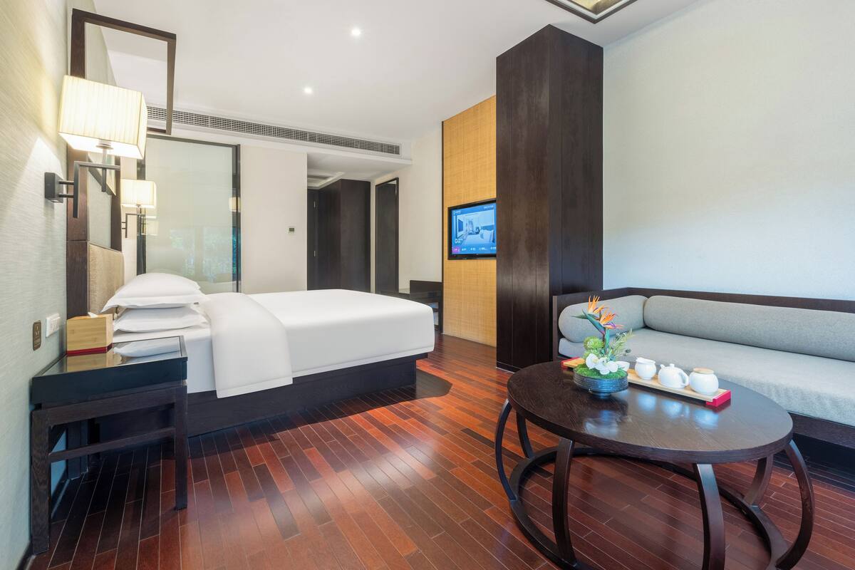 Una habitación de hotel con cama, sofá, una mesita con un arreglo floral y un televisor en la pared.