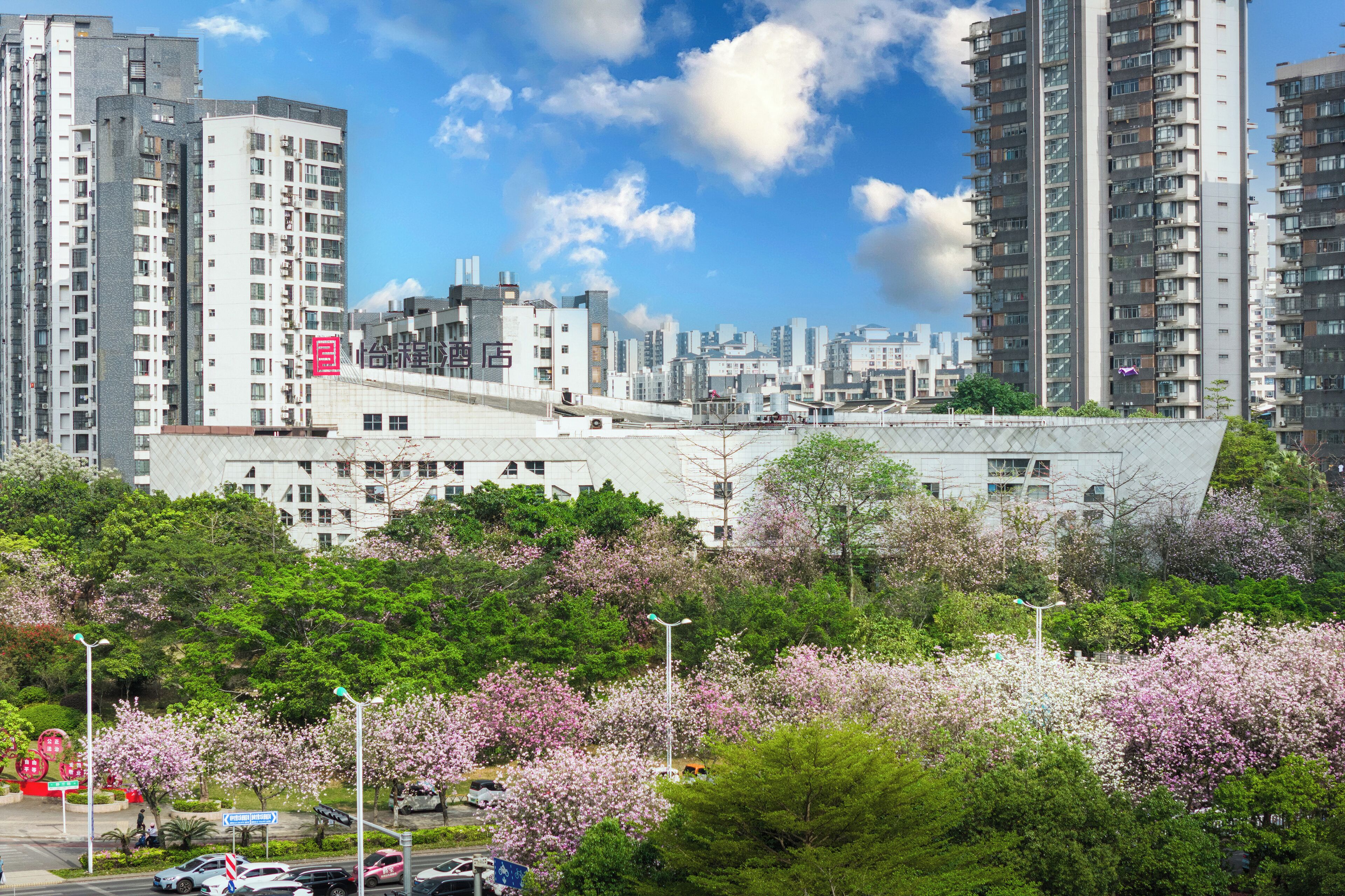 A modern building with a unique angular design, surrounded by lush greenery and flowering trees.