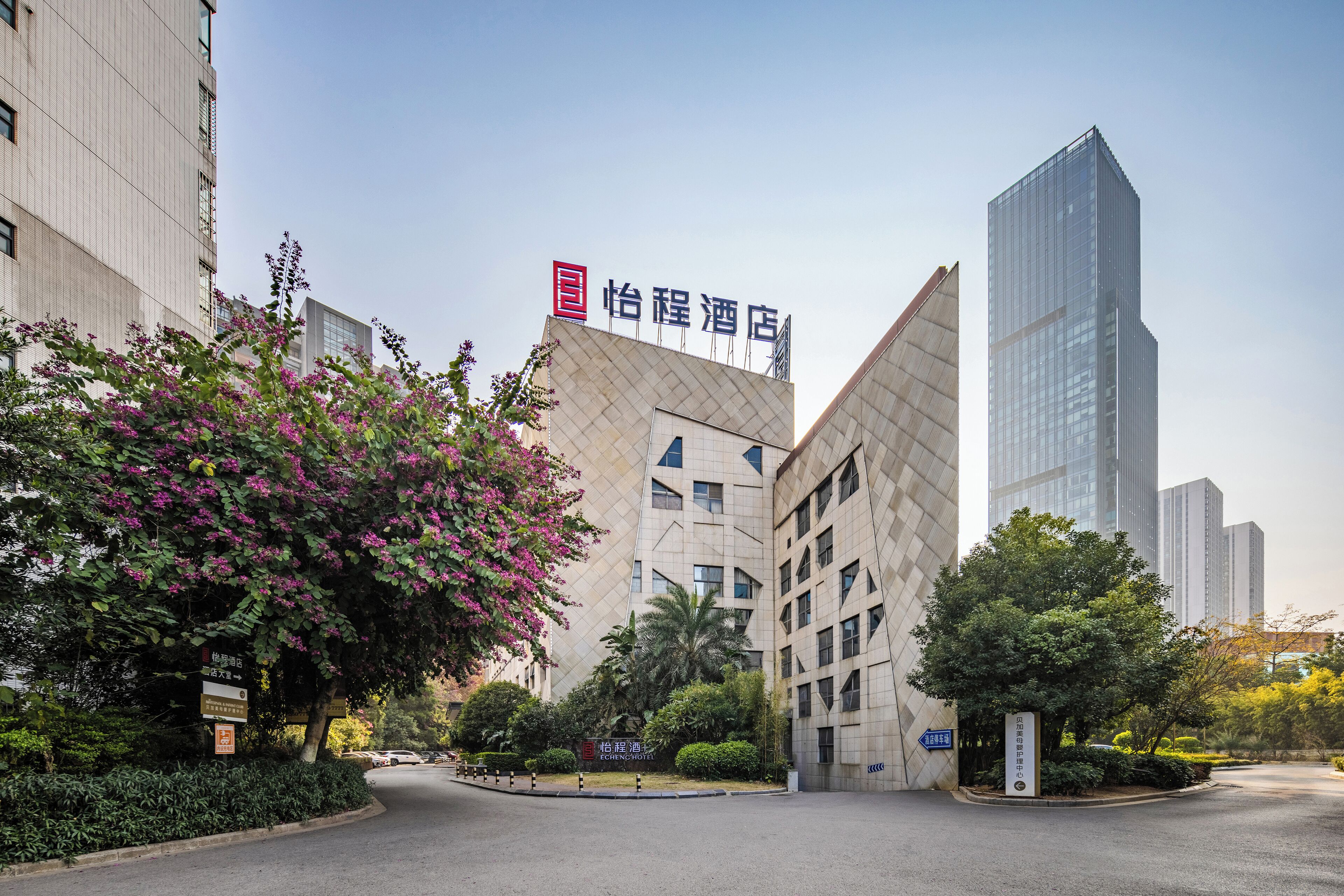 A modern hotel with a distinctive geometric facade, surrounded by lush greenery and adjacent to a tall skyscraper.