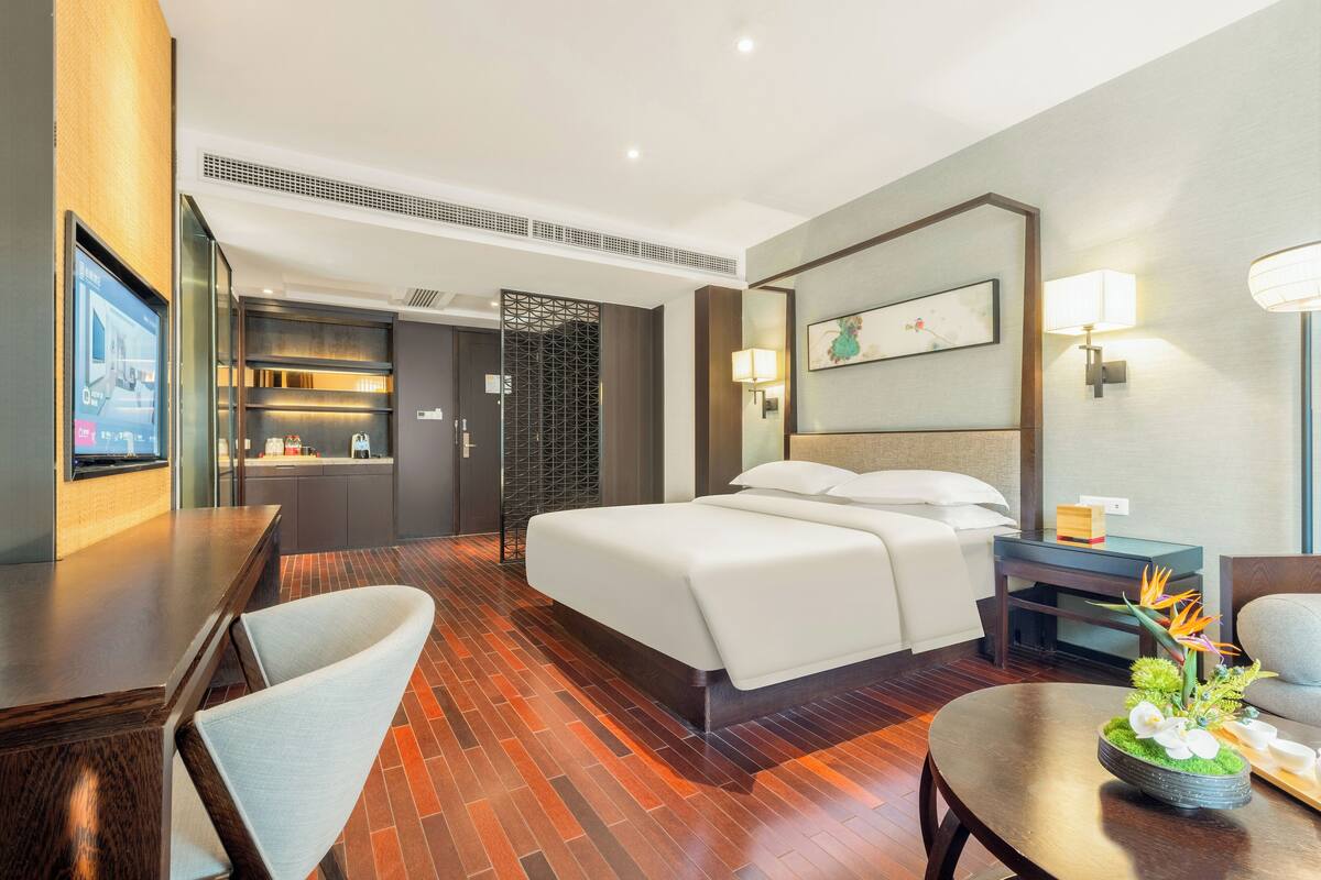 Una habitación de hotel moderna con una cama grande, un escritorio con una silla, una mesita con un arreglo floral y un televisor en la pared izquierda.