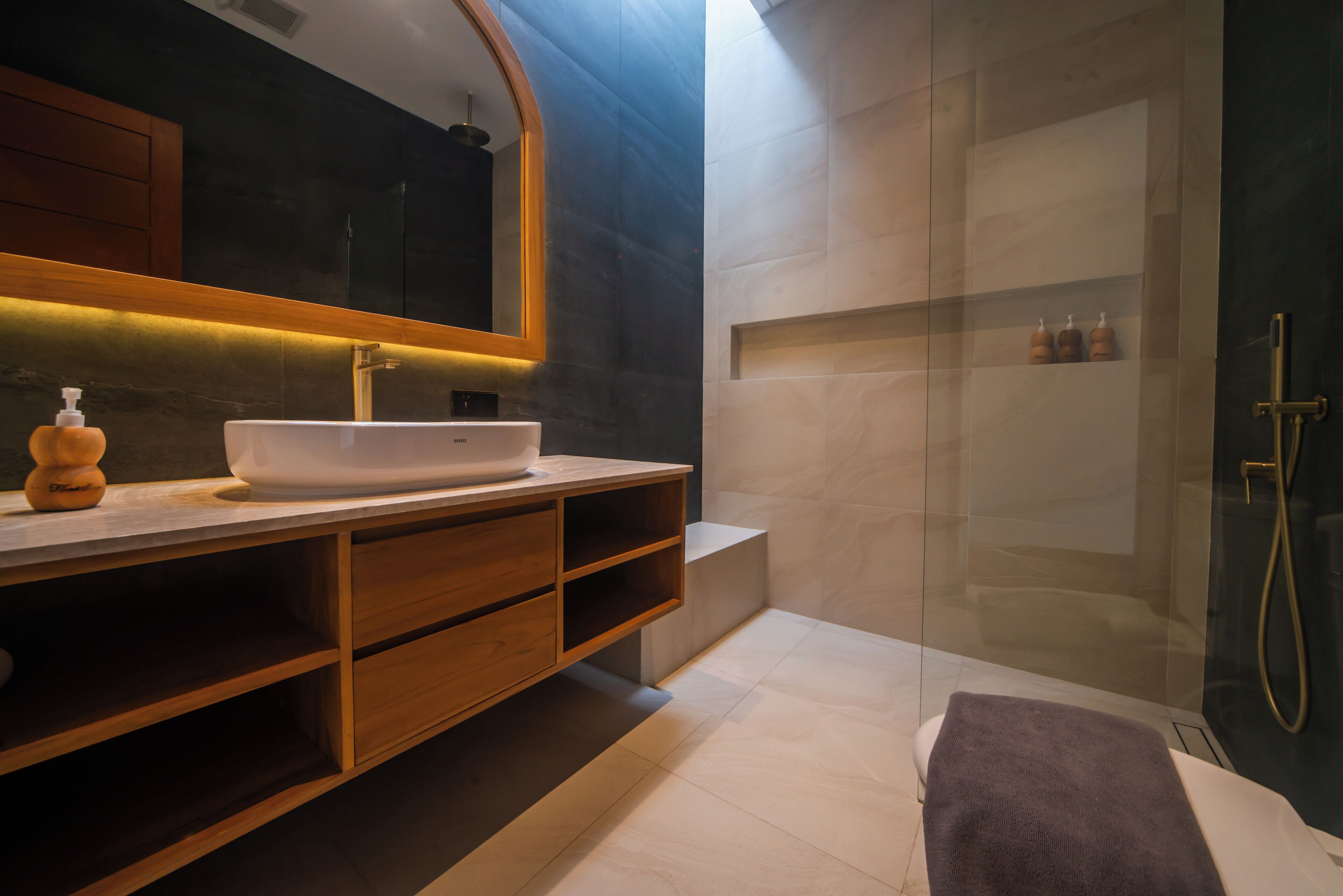 Une salle de bain moderne avec un grand miroir, un lavabo blanc, des meubles en bois et une douche séparée par une paroi de verre.