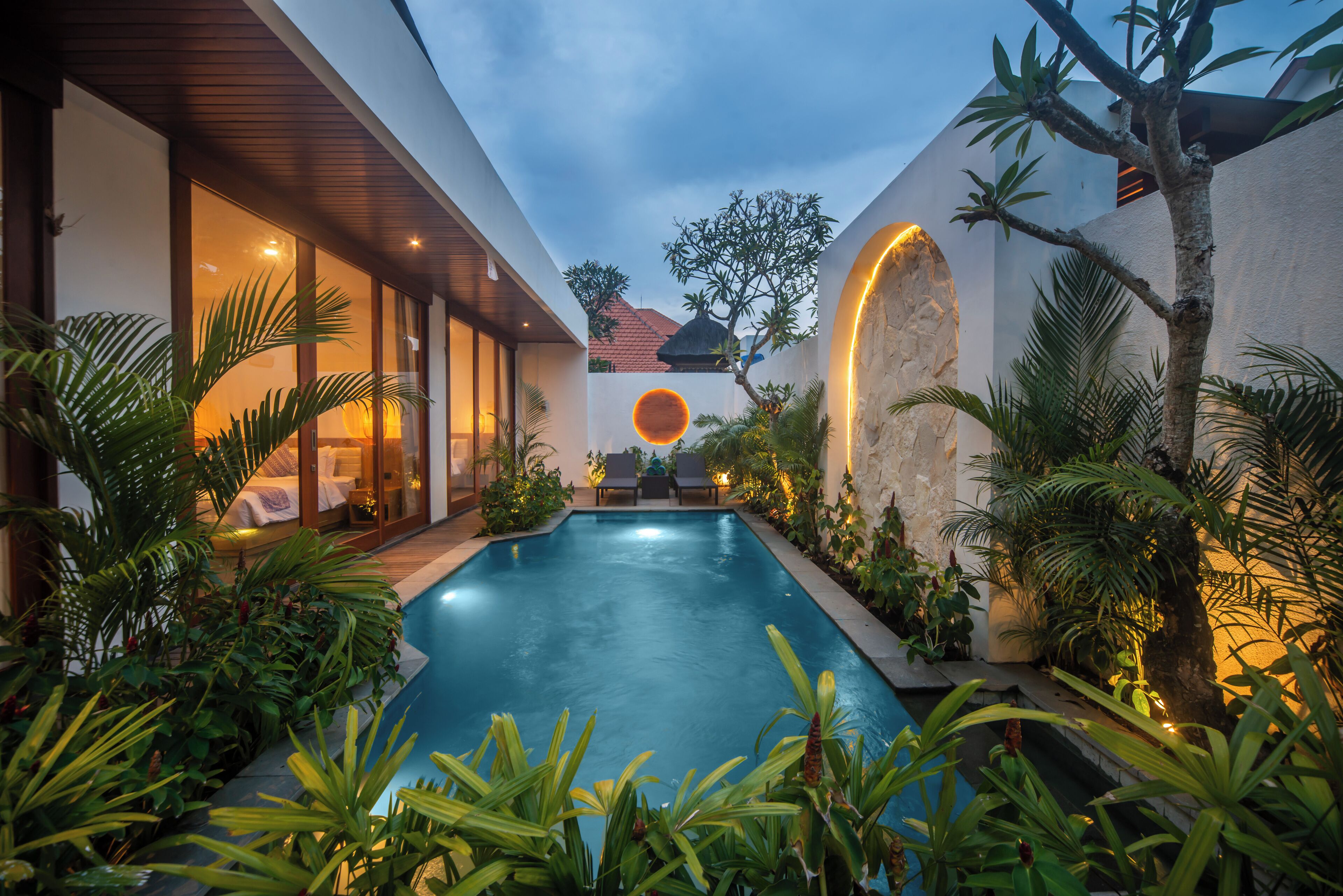 Piscina rodeada por vegetação exuberante e arquitetura moderna, com interior bem iluminado e vista do céu.