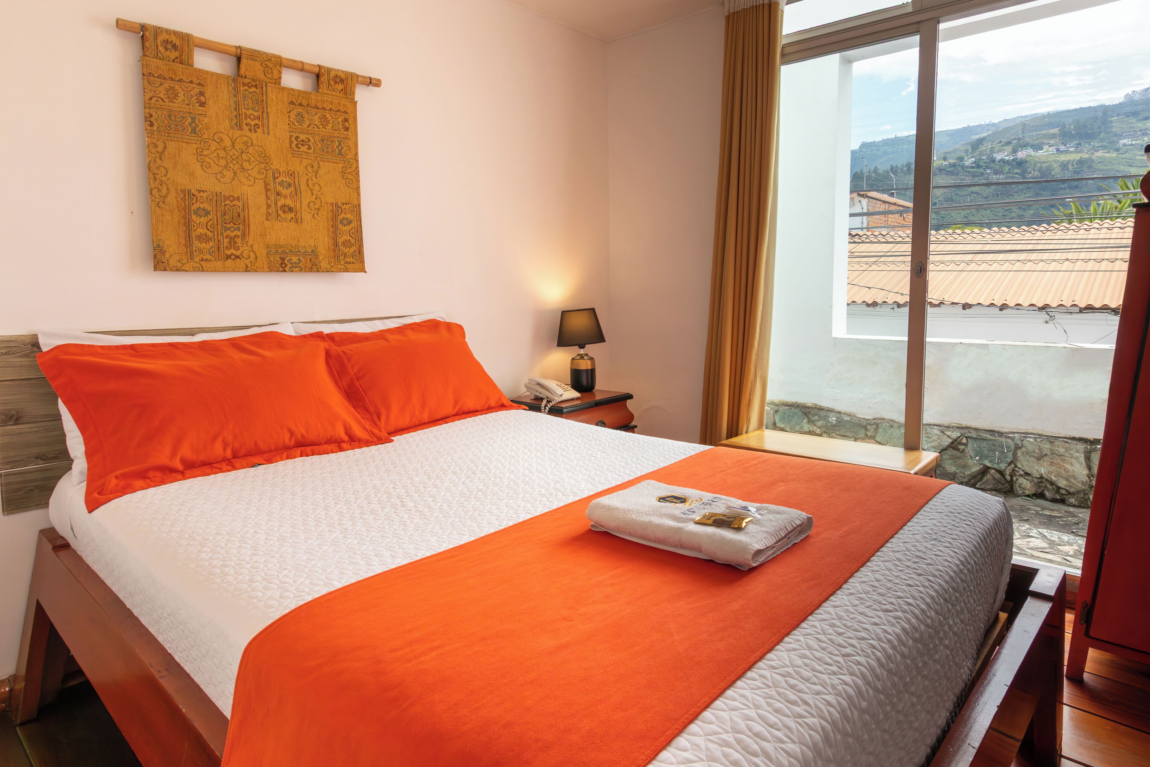 A neatly made bed with an orange and white bedspread, a bedside lamp, and a view of a balcony with a tiled roof.