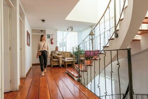 Un amplio salón con suelo de madera, una escalera con barandilla de hierro forjado y un gran ventanal que permite la entrada de luz natural.