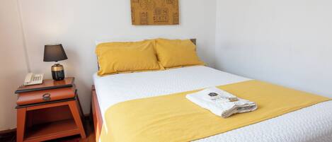 Un letto rifatto con un copriletto bianco e due cuscini gialli, un comodino in legno con una lampada e un telefono, e un tappezzeria decorata sulla parete.