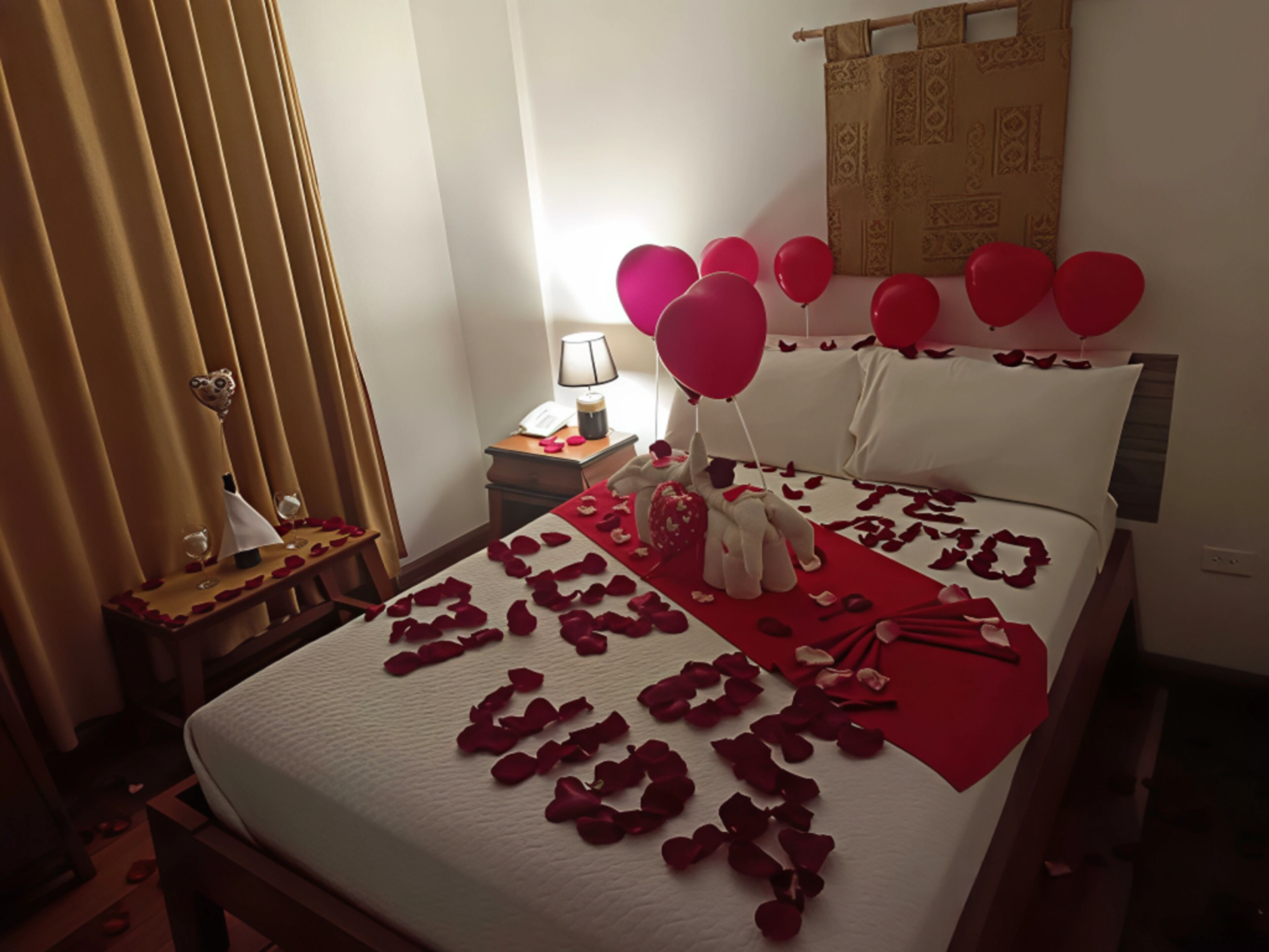 Una camera d'albergo con un letto decorato con petali di rosa e palloncini, un comodino con una lampada e una tenda.