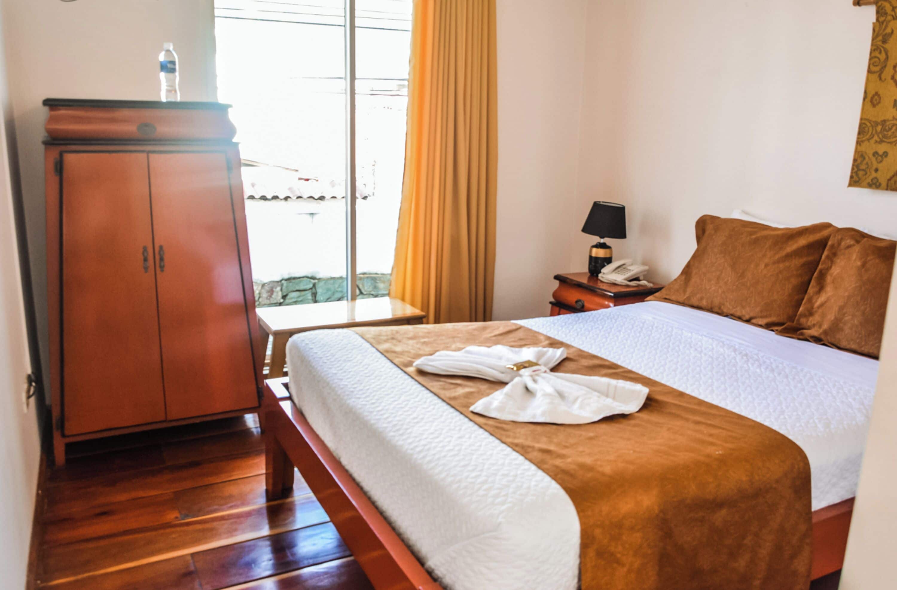 Una habitación de hotel con una cama de madera, una mesita de noche con lámpara, un armario y una ventana con cortinas.