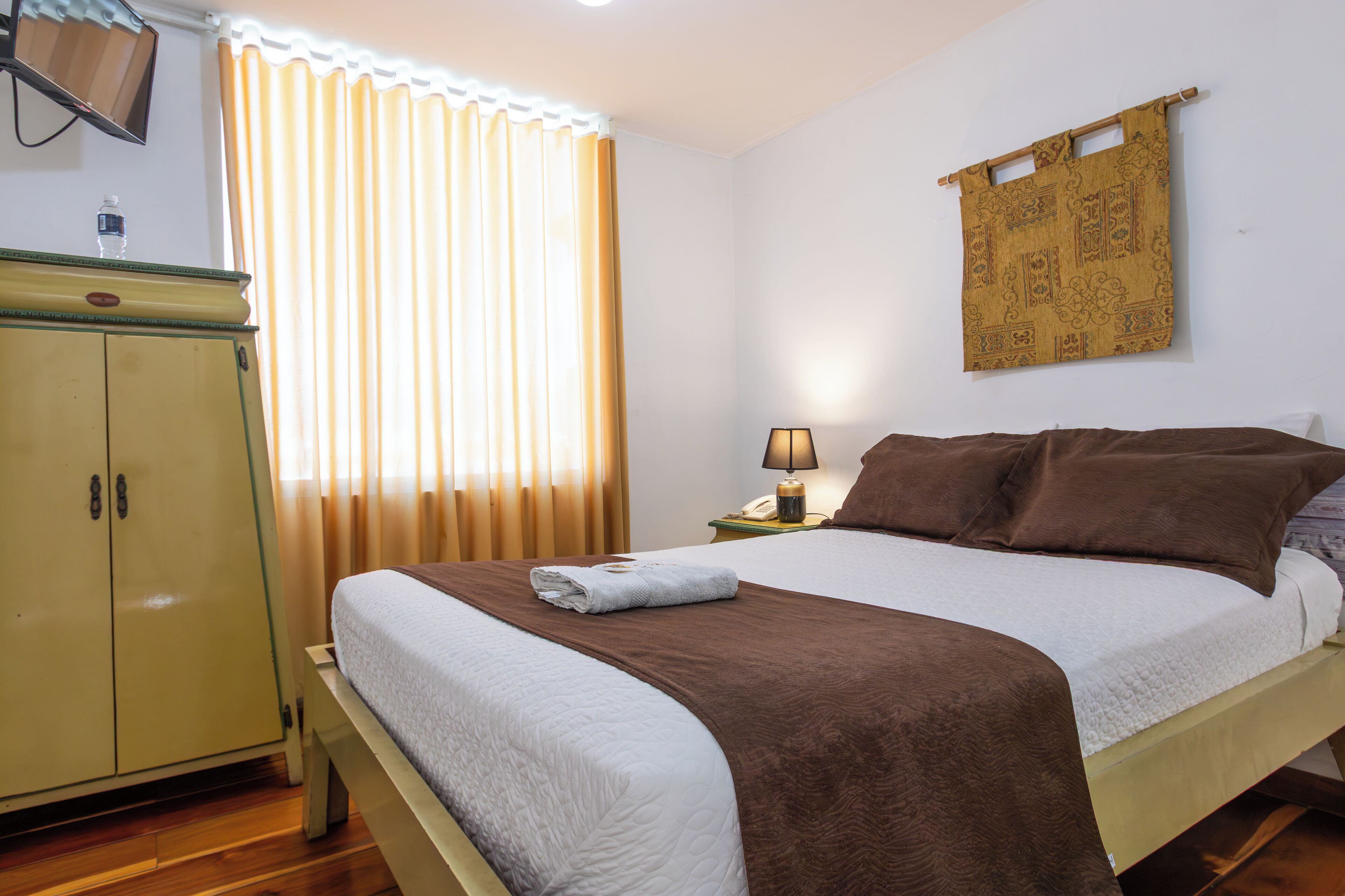 A neatly made bed with a brown blanket, a bedside lamp, and a wall-mounted tapestry.