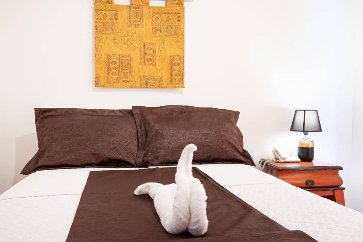 Un lit avec une literie marron, un oreiller blanc et une couverture blanche. Une lampe se trouve sur une table de chevet en bois, à côté du lit.