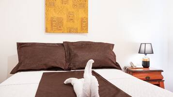 Una cama con ropa de cama marrón, una almohada blanca y una colcha blanca. Hay una lámpara sobre una mesita de noche de madera junto a la cama.