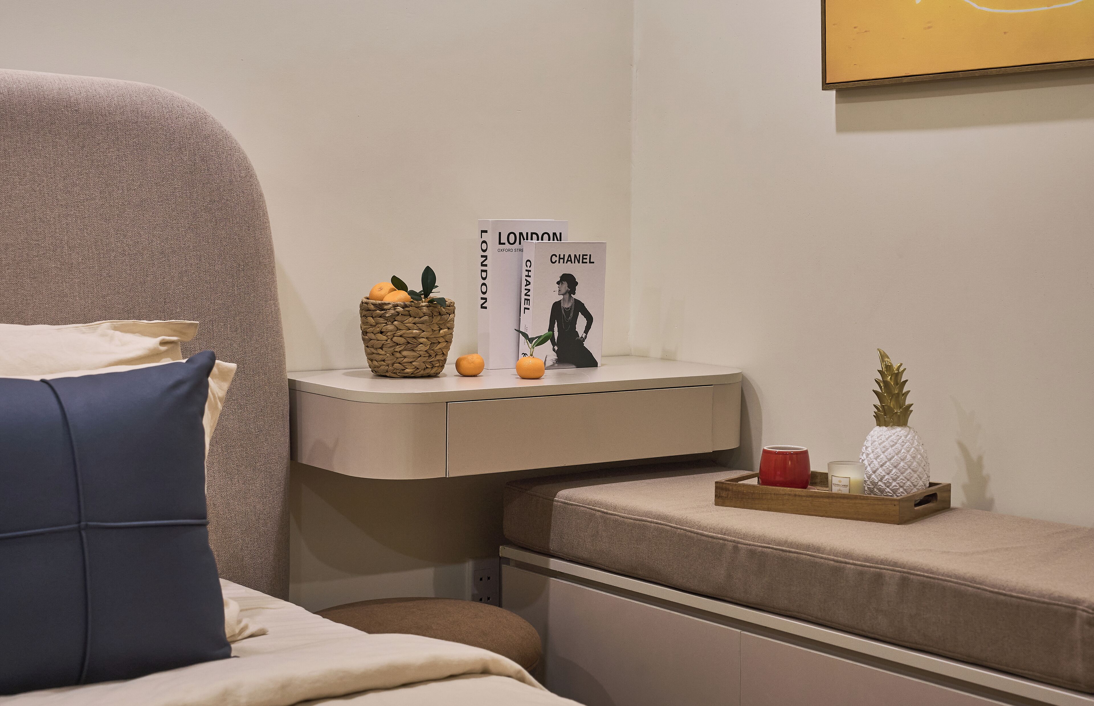 Un dormitorio ordenado con una cama, una mesita de noche con lámpara, una pequeña estantería con libros y objetos decorativos, y un mueble auxiliar con lámpara.