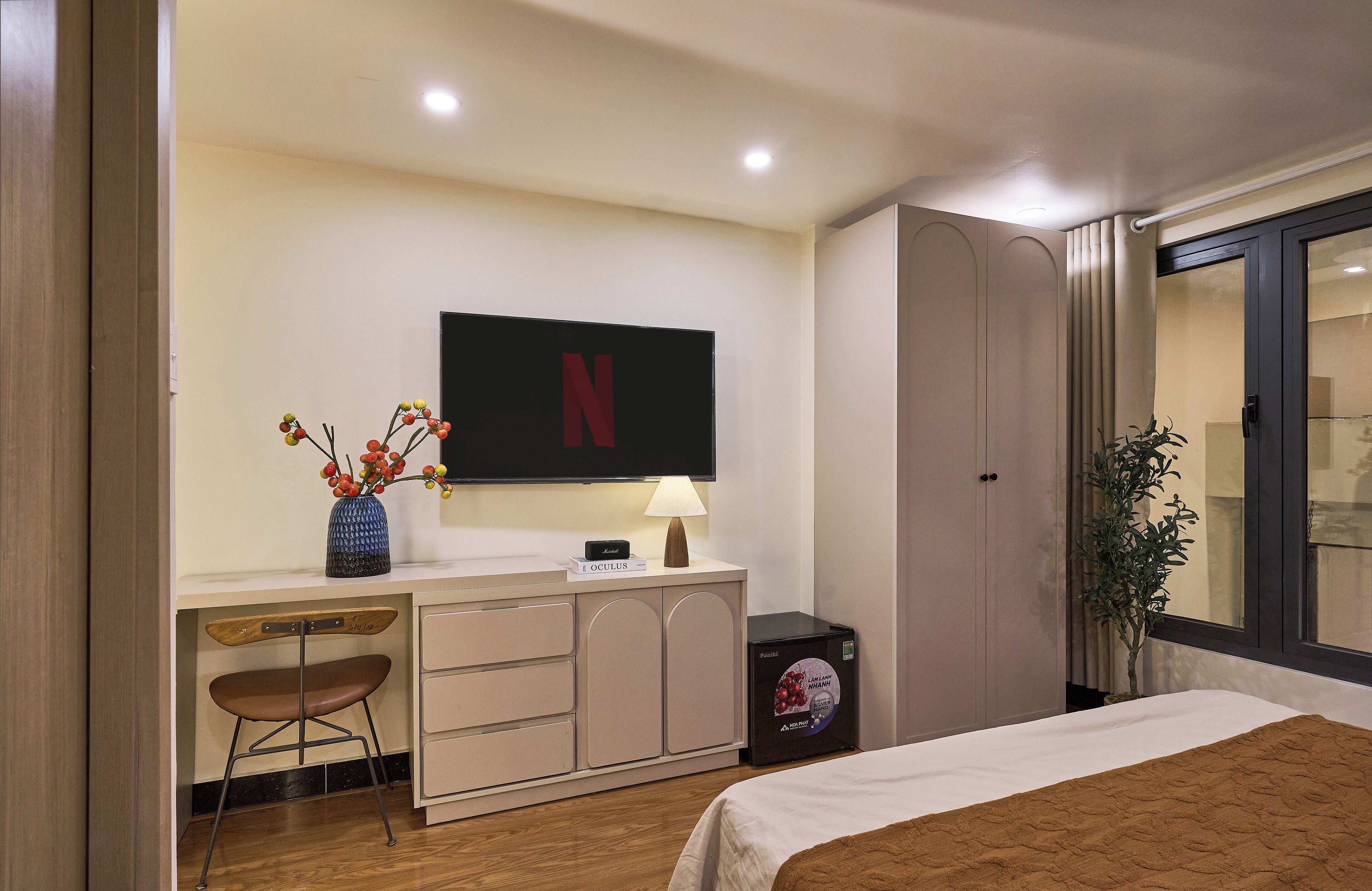 Ein Hotelzimmer mit einem Bett, einem Schreibtisch mit Stuhl, einem an der Wand befestigten Fernseher, einem kleinen Tisch mit Lampe und einer Zimmerpflanze.