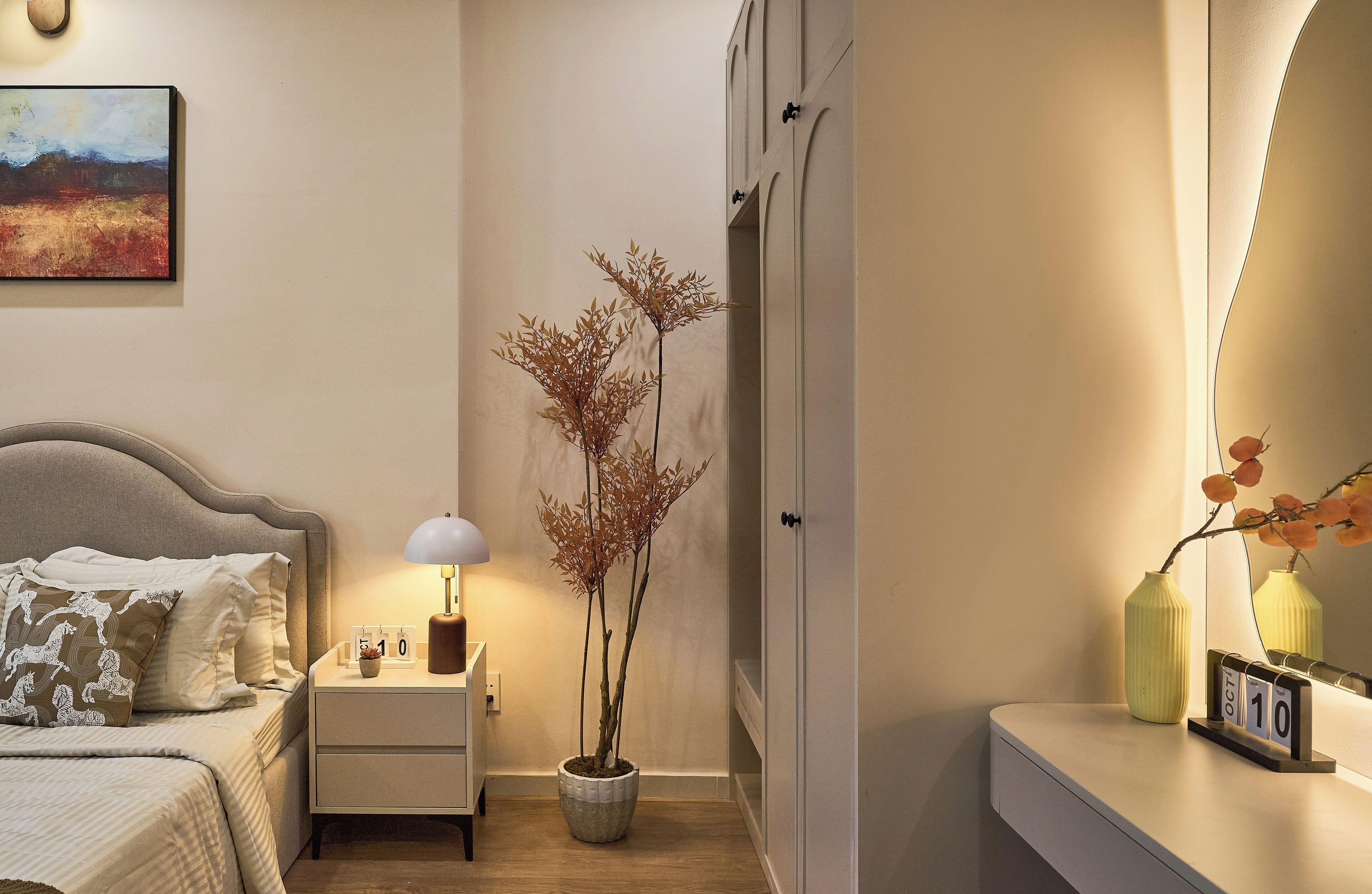 A neatly arranged bedroom with a bed, bedside table, lamp, potted plant, and a mirror reflecting the room.