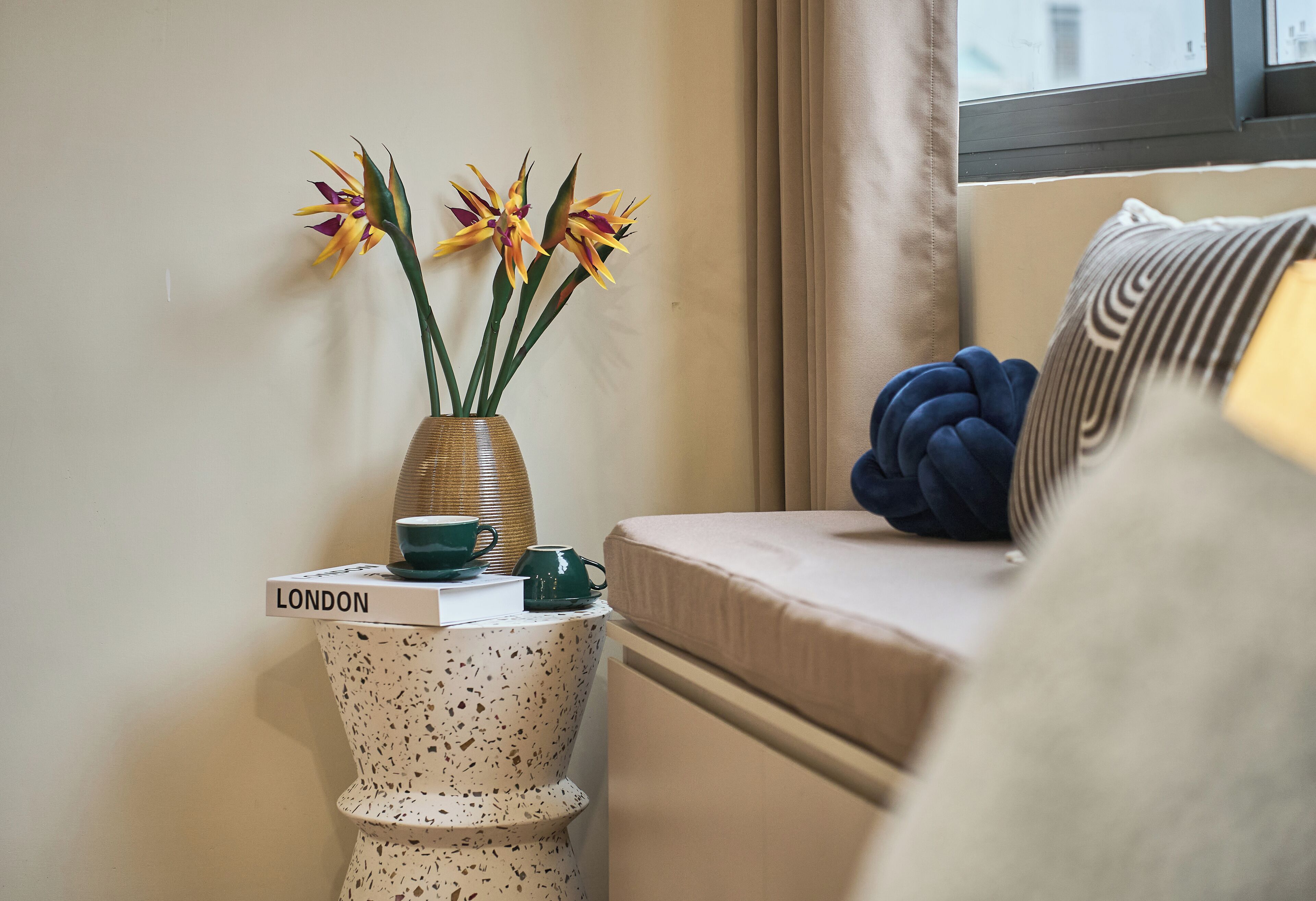 Una habitación acogedora con una cama, una mesita de noche con un jarrón de flores, un libro titulado “LONDON” y una ventana con cortinas.