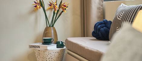 Una habitación acogedora con una cama, una mesita de noche con un jarrón de flores, un libro titulado “LONDON” y una ventana con cortinas.