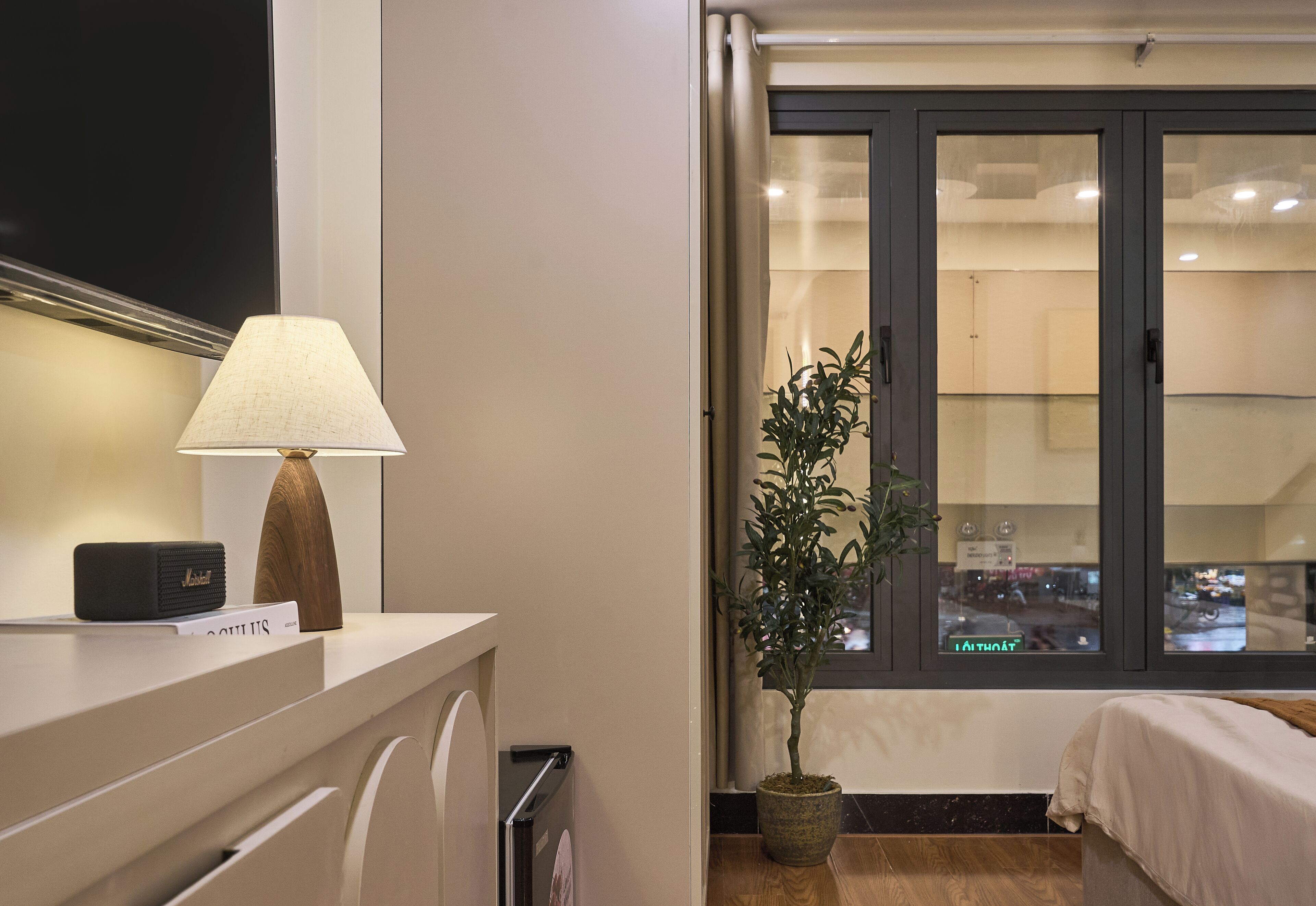 A hotel room with a bed, a bedside table with a lamp, a radio, and a potted plant.