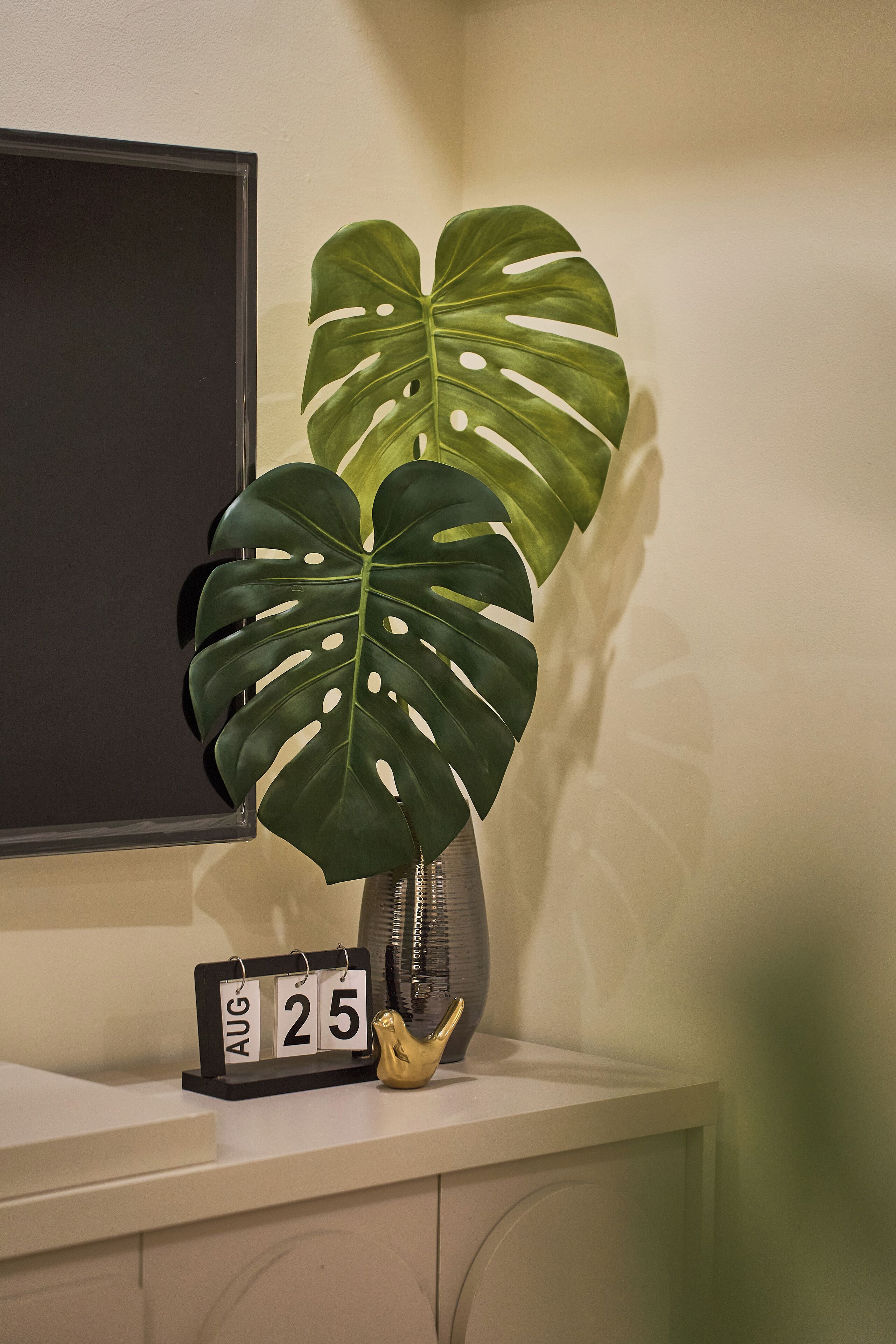 Un calendrier indiquant la date du 25 août, un oiseau décoratif et une grande plante verte posée sur une étagère.