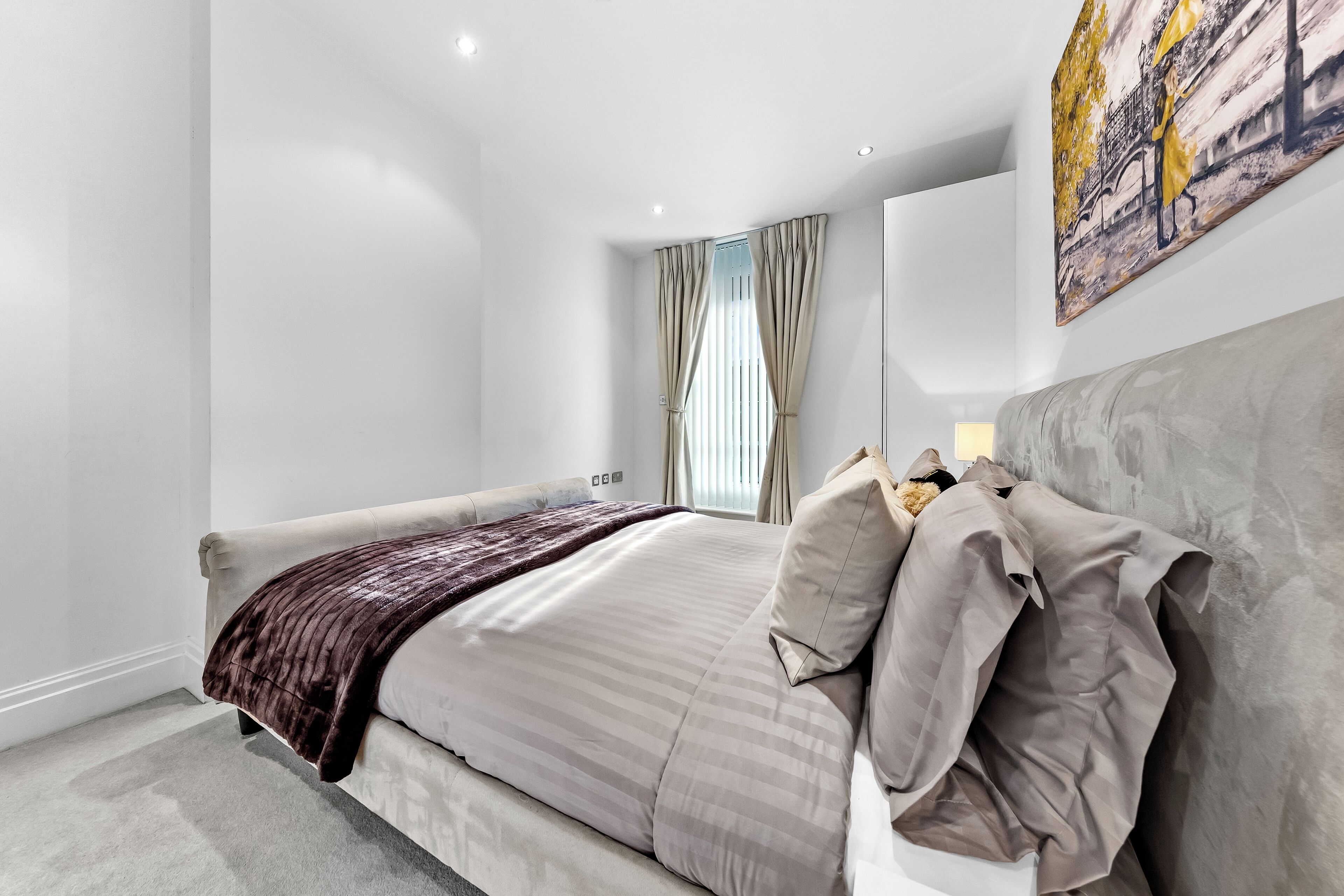 A modern bedroom with a large bed, a headboard, a window with curtains, and a painting on the wall.