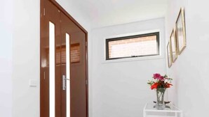 A hallway with a wooden door, a white wall, a small window, and a white table with a vase of flowers.